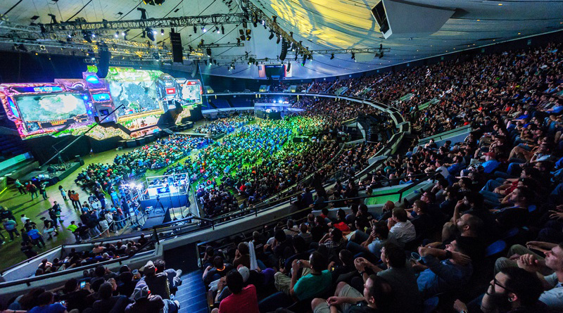 FIFA Esports In Asia: Why do gamers have two headsets? a large stadium packed with people. Green, blue, and yellow strobe lights illuminate the stadium, which focuses on a large screen that shows the esports pro gamers playing a game.